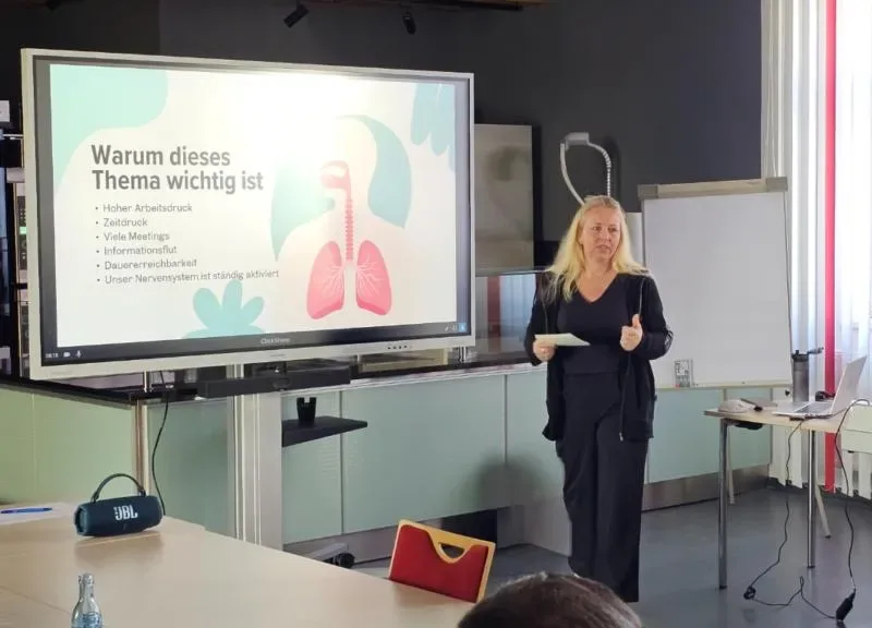 Ines Richter hält einen Atem-Workshop vor Mitarbeitenden, im Hintergrund eine Präsentationsfolie über Stress im Arbeitsalltag